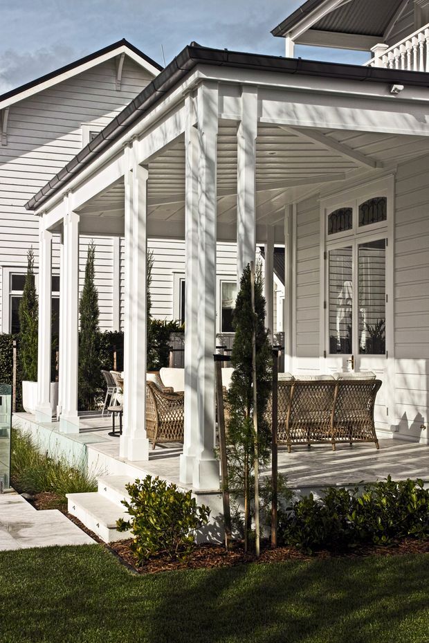 A bright, refined front or side verandah of a large white weatherboard home. The covered outdoor porch has substantial square timber posts, decorative balustrades, a pitched gabled roof, and a timber-lined ceiling. French-style glazed double doors open onto the verandah, which is furnished with wicker seating and surrounded by neatly clipped shrubs and lawn.