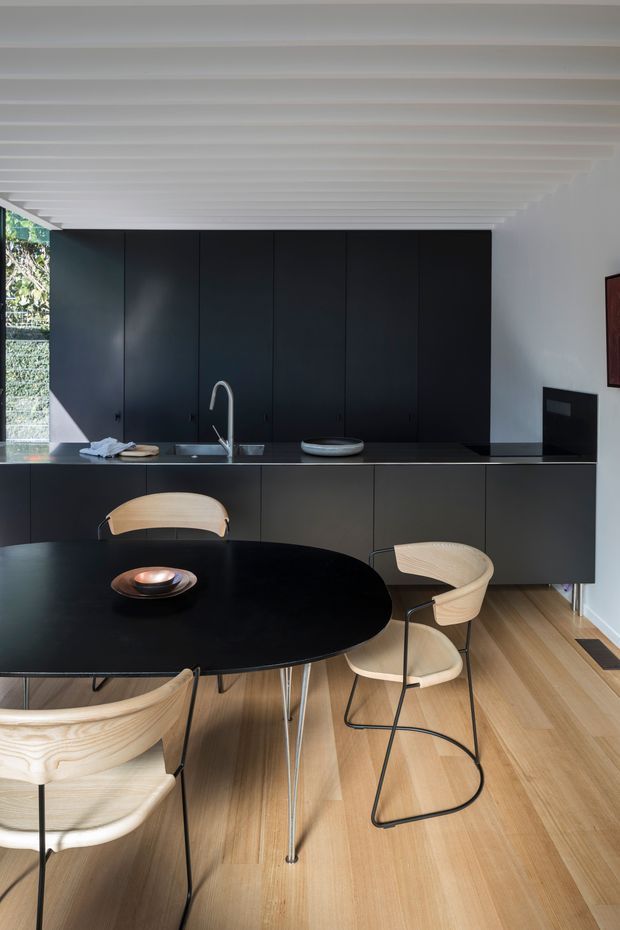 A refined contemporary dining and kitchen interior. A large round black dining table with light timber chairs occupies the foreground, while a wall of matte-black cabinetry and a dark benchtop form the kitchen backdrop. The space has a pale timber floor, white walls, and a distinctive white slatted ceiling with strong linear repetition. Natural light enters from glazing at the left, creating a calm, high-contrast monochromatic scheme softened by timber furniture and flooring.