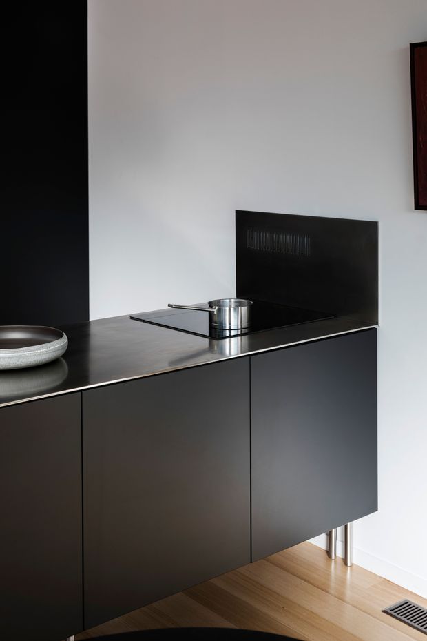 A minimalist contemporary kitchen vignette featuring matte dark cabinetry, a dark countertop with integrated black cooktop, stainless steel saucepan, and matching dark splashback. The cabinetry is elevated on slim metal legs above a warm timber floor, with a mostly white wall and a linear floor vent visible.