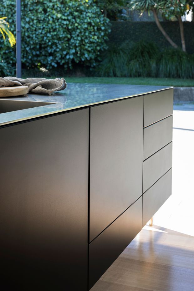 Close-up architectural photograph of a sleek, minimalist kitchen island with flush-panel dark taupe cabinetry, thin black reveals, and a pale blue-grey stone benchtop. The island sits beside large glazing or an open connection to a lush landscaped garden and pool area, with warm timber flooring and strong natural light.