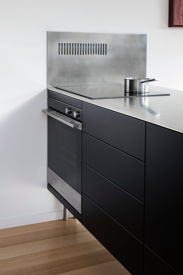 Close-up view of a compact contemporary kitchen with matte black handleless cabinetry, an integrated stainless-steel oven, stainless-steel benchtop and splashback, and a black glass cooktop. Light timber flooring and white walls provide contrast. The splashback includes a narrow horizontal ventilation grille, and a small stainless-steel saucepan sits on the cooktop.