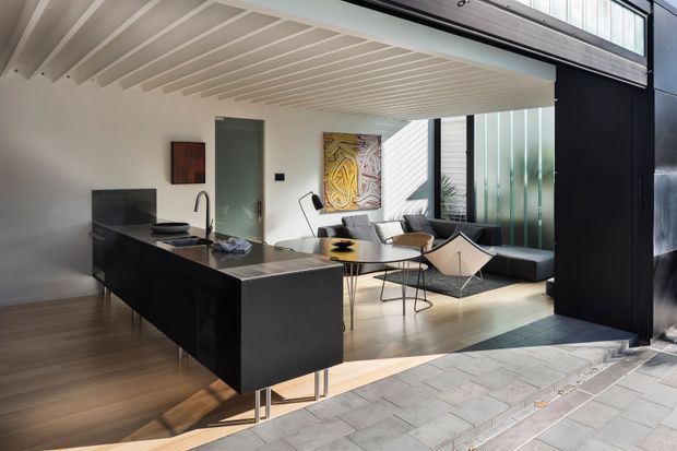 A contemporary open-plan interior viewed from an exterior threshold, combining a compact black kitchen island with an integrated sink and cooktop, a dining area, and a relaxed living zone. The space features warm timber flooring, white walls, a white slatted ceiling, dark built-in cabinetry, expansive glazing, and a strong connection to the paved outdoor area. Sculptural mid-century-inspired furniture and artwork add character, while abundant daylight creates a bright, refined atmosphere.