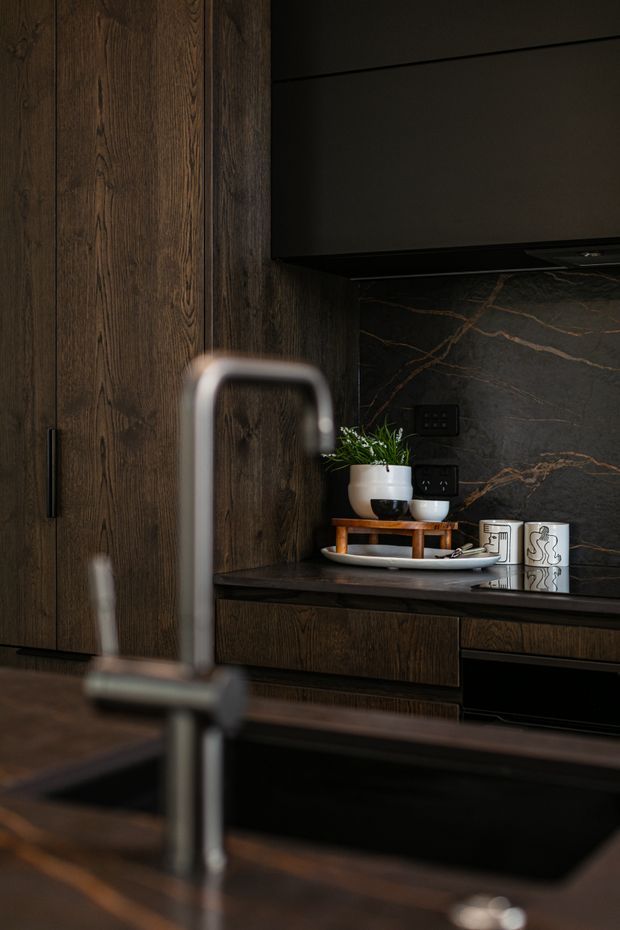 A moody, sophisticated kitchen interior with dark stained timber cabinetry and wall panelling, a black stone island benchtop with integrated sink and tall brushed-metal tap, and a dramatic dark veined stone splashback. Black overhead cabinetry, dark electrical fittings, and understated decorative ceramics reinforce the refined palette.