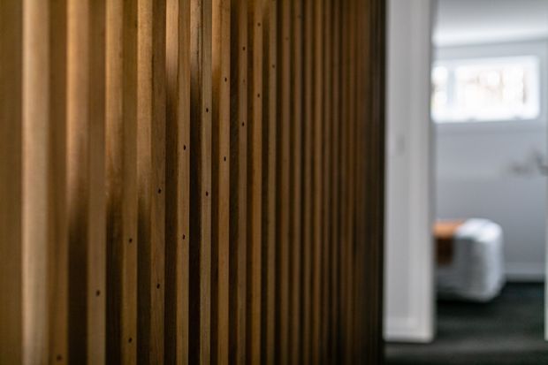 Close-up interior view of a dark stained timber feature wall or vertical slatted partition, with a softly blurred adjoining room visible through an opening. The space has a warm, tactile material palette and subdued lighting.