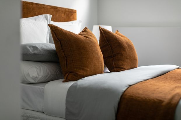Close-up view of a neatly made bed in a bedroom, featuring crisp white bedding, layered white pillows, rich rust-brown velvet cushions, a matching rust-brown throw, and a warm timber headboard against a light neutral wall. The styling is simple, warm, and refined.