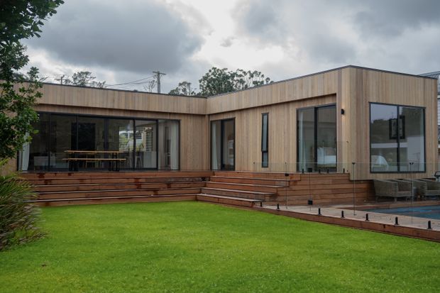 A contemporary single-storey residence with a low-profile flat roof and a warm timber-clad facade. Large glazed openings connect the interior to a broad timber deck and stepped outdoor entertaining area, overlooking a manicured lawn and a partially visible pool. The image is photographed from the garden under an overcast sky.