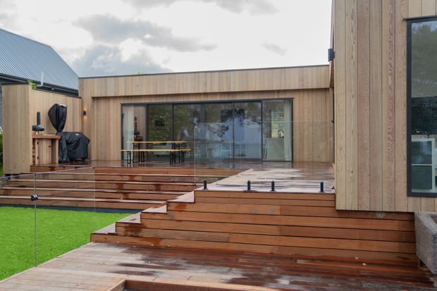 Exterior view of a contemporary timber-clad home arranged around a stepped timber deck and courtyard-like outdoor living area. Broad wet timber stairs, glass balustrades, large sliding glazed doors, and a low flat-roofed pavilion are visible, with an adjacent dark pitched roof volume in the background.