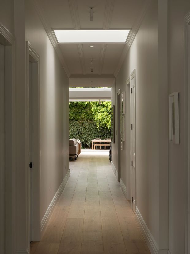 Long, light-filled residential hallway with pale timber flooring, white walls, detailed cornices and skirting, multiple white-panelled doorways, and a large rectangular skylight. The corridor opens toward a bright living area and leafy outdoor courtyard, creating a strong indoor-outdoor connection.