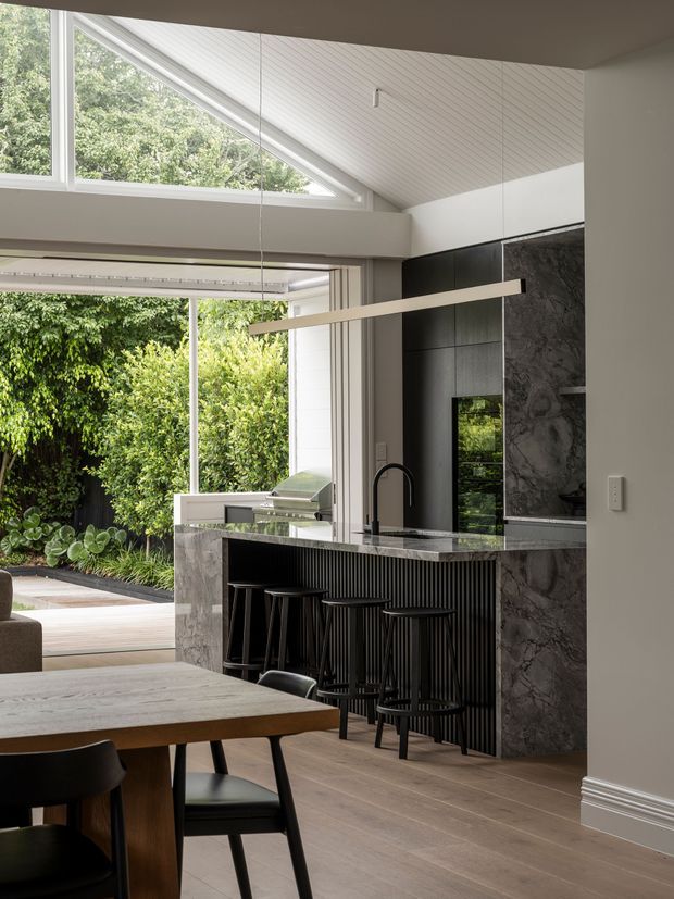 A refined open-plan kitchen and dining area with a dramatic vaulted, white-lined ceiling and extensive glazing opening to a leafy outdoor space. The kitchen features a dark fluted island with a pale veined stone benchtop, matching dark cabinetry, a large stone feature wall, integrated appliances, and black stools. A timber dining table and black chairs occupy the foreground, while an outdoor grill is visible beyond the open glazing. The space has a sophisticated contemporary character with strong indoor-outdoor connection and abundant natural light.