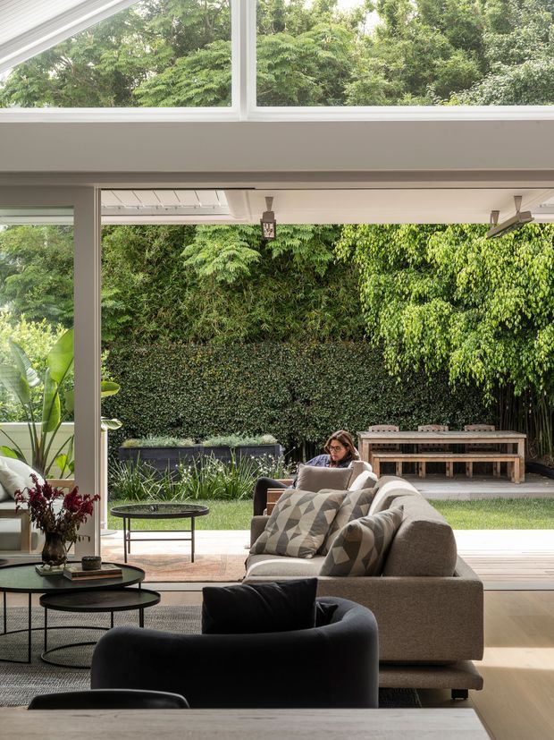 A bright, contemporary living room opens directly onto a lush private garden through expansive floor-to-ceiling glazing and wide sliding doors. The room features a large neutral sectional sofa, black lounge chairs, layered rugs, round coffee tables, and abundant indoor plants. The garden includes dense green screening, a paved entertaining area, a raised planter or water feature, and a long timber outdoor bench. A high glazed clerestory brings additional daylight into the space.