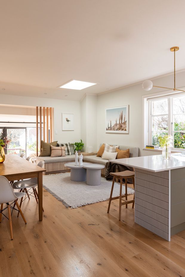 Bright open-plan residential interior combining a living room, dining area and kitchen. The space features warm timber flooring, a pale neutral ceiling and walls, a grey sofa with layered cushions, a timber dining table, pale upholstered chairs, and a kitchen island clad in small light tiles with a white benchtop. Vertical timber battens partially screen the living area, while large windows provide abundant natural light. The overall styling is warm, contemporary and refined.