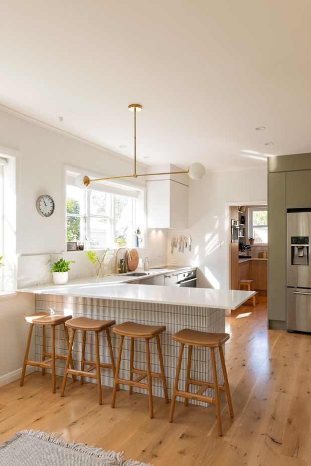 Bright, spacious contemporary kitchen with a large white island and integrated breakfast bar, vertical fluted tile detailing, light timber flooring, white cabinetry and stone surfaces. Four timber bar stools line the island, while a sculptural brass-and-glass pendant light hangs overhead. Large windows provide abundant natural light, and a stainless steel refrigerator is integrated into tall cabinetry at the right.