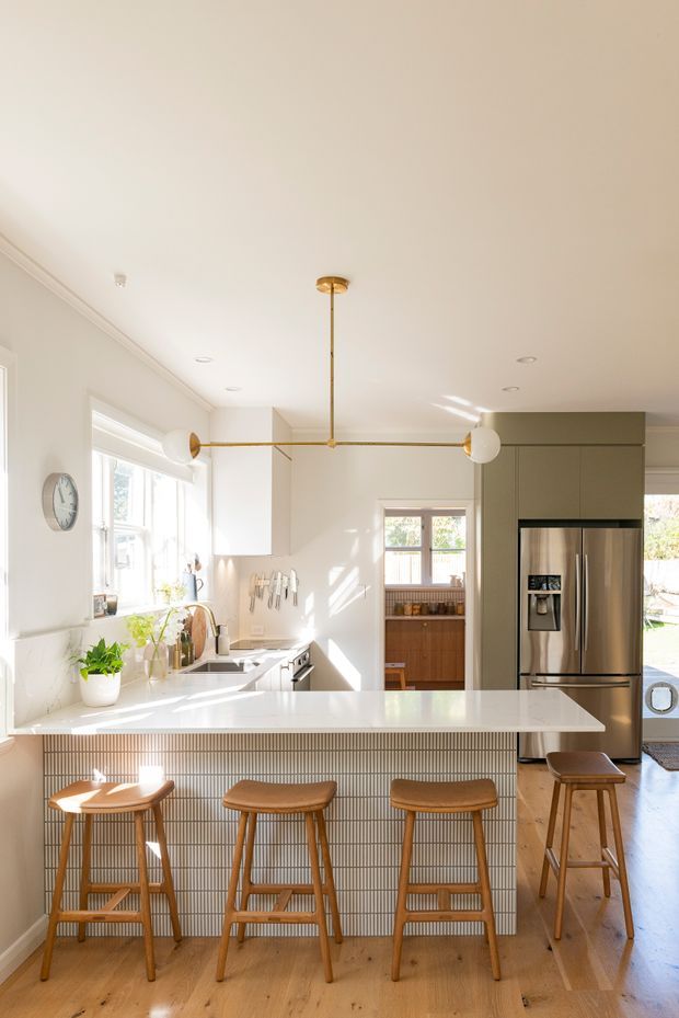 Bright, warm contemporary kitchen with a large white island and integrated breakfast bar, vertical light-toned tiled island face, timber bar stools, pale timber flooring, white cabinetry, stainless-steel refrigerator, and brass globe pendant lighting. Sunlight enters through large windows, creating an airy residential interior.