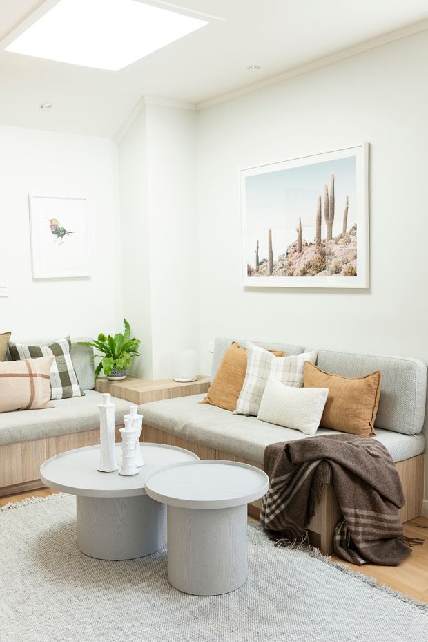 Bright, serene living room with pale upholstered built-in seating, warm timber bases and flooring, layered neutral cushions, twin round coffee tables, a textured rug, indoor greenery, framed artwork, and a large ceiling skylight.