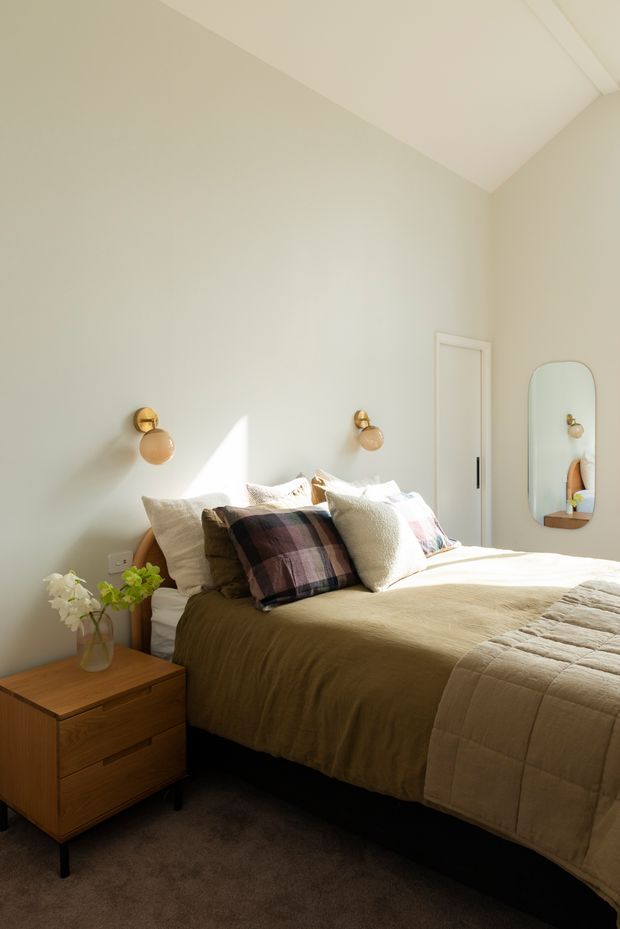 A warm, softly lit bedroom with a low bed dressed in neutral beige bedding and layered textured pillows, including a plaid accent cushion. The room features pale green-grey walls, a timber bedside table with drawers, brass-and-glass wall sconces, a full-height white door, an arched wall mirror, and dark carpet flooring. Sunlight creates a bright triangular highlight across the wall and bed.