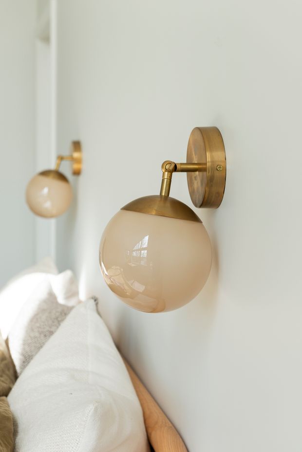 Close-up interior photograph of a brass wall-mounted sconce with a rounded, glossy cream glass globe beside a bed. A second matching sconce and soft white bedding are visible in the background, creating a warm, refined bedroom setting.
