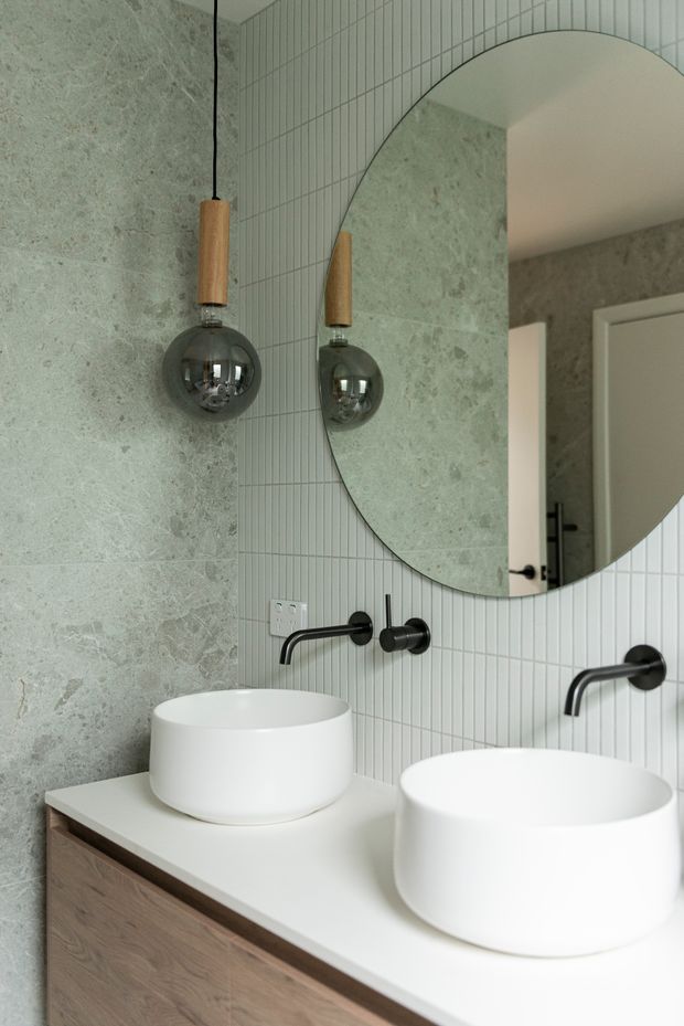 A contemporary bathroom vanity with two white vessel basins, a pale timber cabinet, matte black wall-mounted tapware, a large rounded mirror, pale green-grey stone wall tiles, and narrow white vertical tiles. Pendant lights with timber details and smoked glass globes hang beside the mirror.