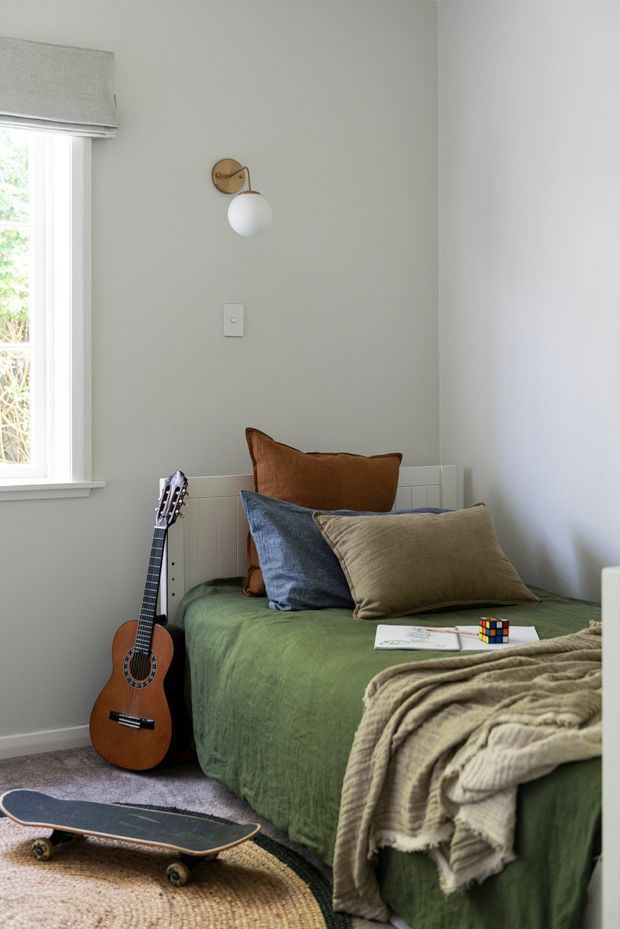 A cosy, compact bedroom with a single bed dressed in a muted green cover, layered blue, tan and brown cushions, and a textured beige throw. A timber acoustic guitar rests beside the bed, with a skateboard in the foreground. The room has pale grey walls, white trim, a small window with a Roman blind, a simple wall-mounted globe sconce, a woven round rug and a playful Rubik’s cube on the bed. The styling feels warm, relaxed and youthful.