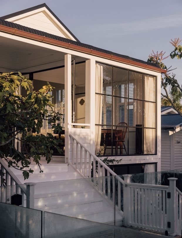 Exterior view of a raised, light-painted timber house with a gabled roof and corrugated dark roofing. The facade features a covered porch, enclosed sunroom or living area with large black-framed glazing and curtains, white balustrades, stairs, and a glass-and-white fence. Mature planting partially screens the front elevation.