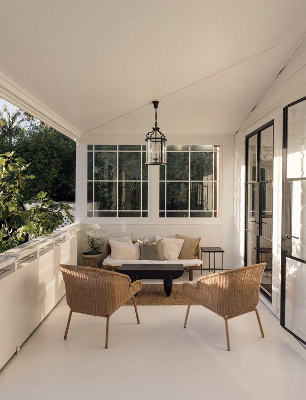A bright enclosed veranda or sunroom furnished as a relaxed sitting area. The space has white paneled walls and ceiling, large multi-pane windows, black-framed glazed doors, a woven rattan sofa and two matching armchairs, a small dark coffee table, and a hanging lantern-style pendant. Lush greenery is visible outside, creating an airy indoor-outdoor atmosphere.