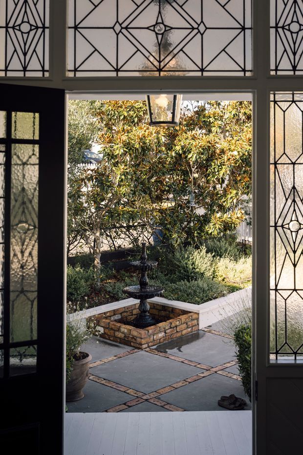 View from inside through an open entry door into a landscaped courtyard. The foreground features a dark-painted timber threshold and decorative leaded-glass sidelights and transom with geometric diamond and circular motifs. Outside is a paved courtyard with intersecting brick borders, a small tiered black fountain set in a low brick planter, and established shrubs and trees.