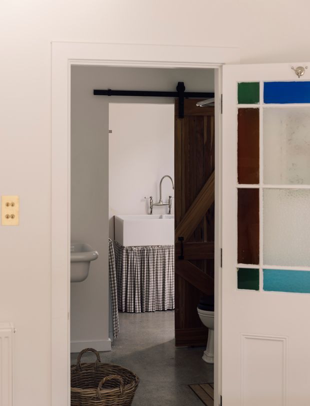 View from a pale, traditional interior hallway through an open doorway into a compact bathroom or laundry area. The room contains a white ceramic utility sink with a curved chrome tap, checked fabric skirting, a partially visible toilet, and a grey floor. A dark timber barn-style sliding door with black hardware is open beside the doorway. The foreground door has decorative coloured and textured glass panels in green, blue, brown, and turquoise, with a white raised-panel lower section. A wicker basket sits on the floor.