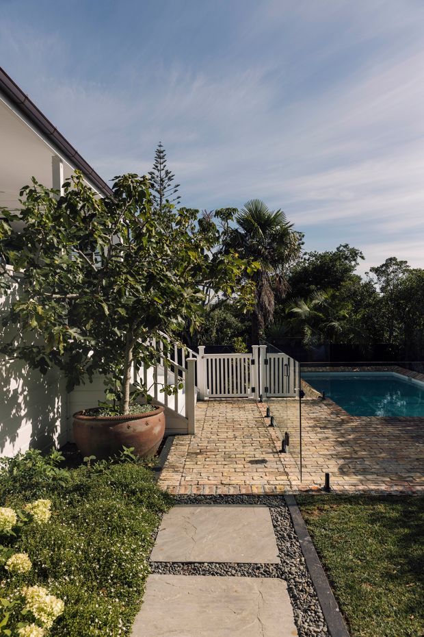 A landscaped residential backyard viewed from a stone stepping-stone path. The scene features a white weatherboard-style house with a sloping roof edge, lush tropical and subtropical planting, a large terracotta planter, white picket-style pool gate and balustrade, and a rectangular swimming pool bordered by warm brick paving. The outdoor space is sunny and established, with a relaxed garden setting and strong indoor-outdoor connection.