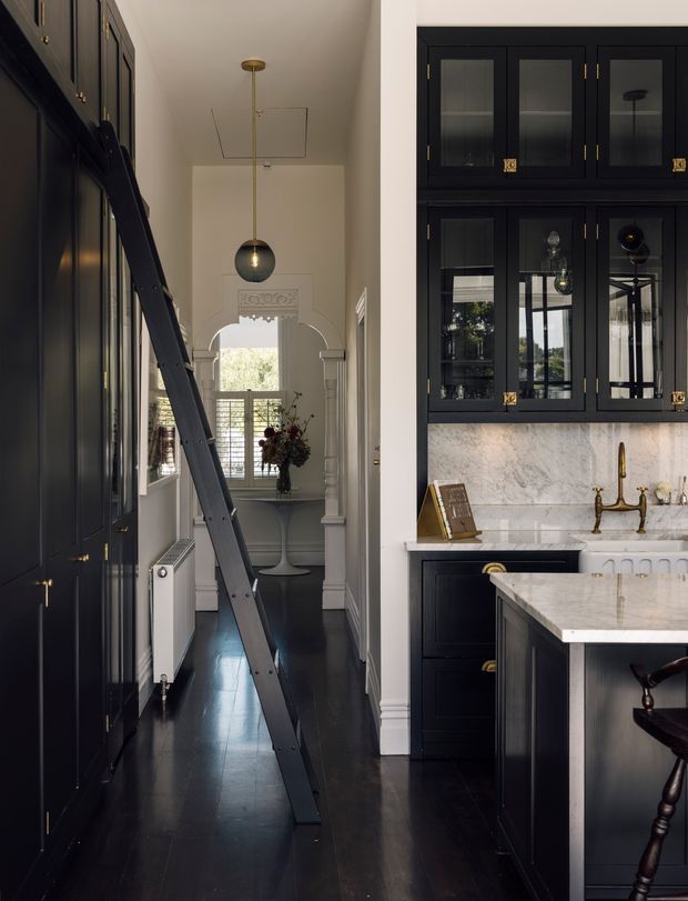 A narrow, sophisticated kitchen and hallway interior with dark navy-black full-height cabinetry, glazed display cupboards, brass hardware, dark timber flooring, and a pale marble backsplash and island benchtop. A ladder leans against the cabinetry, while the hallway beyond features an ornate arched opening, pendant lighting, a small console table, and a balcony or window at the far end. The scheme combines traditional detailing with a refined contemporary kitchen fit-out.