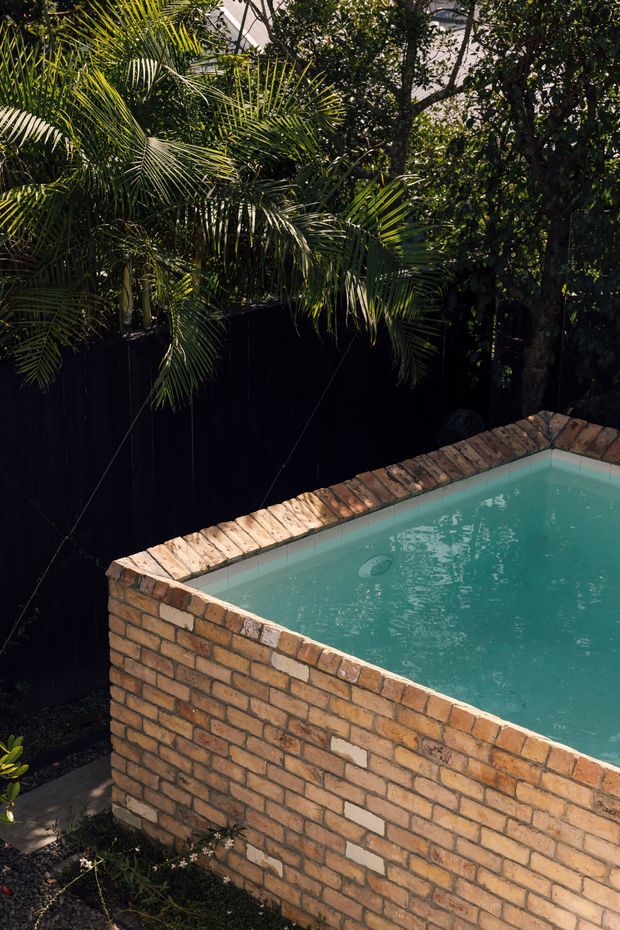 A close-up outdoor view of a raised rectangular swimming pool with turquoise water, built into a warm exposed-brick structure. The pool edge has brick coping, and dense tropical palms and leafy planting surround a dark privacy fence, creating a secluded garden setting.