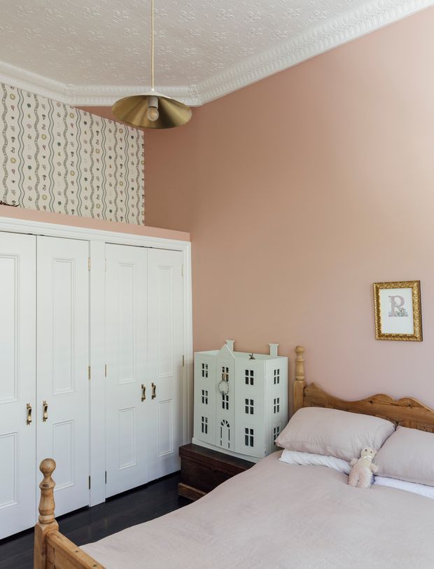 A charming traditional bedroom with a blush pink painted wall, ornate white embossed ceiling and decorative crown moulding. The room features a large white paneled built-in wardrobe with brass hardware, dark timber flooring, a warm timber bed with turned posts and carved detailing, pale bedding, and a small white dollhouse-style cabinet. A patterned wallpaper panel and brass pendant light add vintage character.