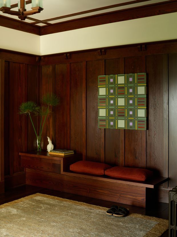 A richly detailed traditional interior entry or hallway featuring dark stained timber wall panelling, exposed timber beams and decorative ceiling trim. A built-in timber bench with rust-red cushions sits beneath a geometric abstract artwork, beside a narrow console displaying books and a vase. A neutral woven rug and warm low lighting create a moody, refined atmosphere.