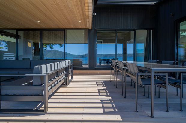 A spacious modern outdoor terrace or covered deck with timber decking, a long dining table and chairs, and built-in outdoor seating. Dark vertical wall cladding and a warm timber ceiling frame extensive glazing, with views of blue water and distant hills visible beyond. Strong linear shadows create a high-contrast, open and refined atmosphere.
