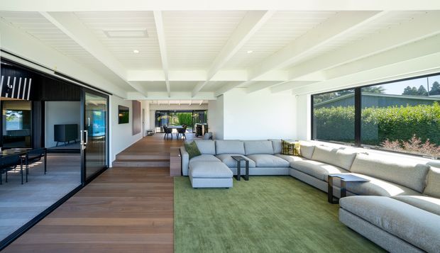 Bright, spacious open-plan living room with a large modular sectional sofa and green area rug, connected to a dining area through a wide stepped transition. The room features expansive dark-framed glazing, timber flooring, white walls and a white painted exposed-beam/coffered ceiling. A dark-framed glazed opening on the left reveals an adjacent room or workspace. The interior has a clean contemporary feel with abundant natural light and garden views.