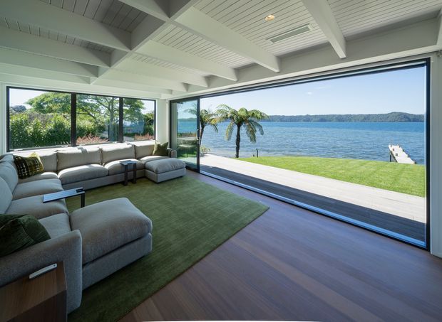 Bright waterfront living room with expansive corner glazing and a wide opening to a timber deck and lawn overlooking a calm body of water. The room features a large pale sectional sofa, green area rug, dark timber flooring, and a white painted exposed-beam and tongue-and-groove ceiling. A small dock and palm trees are visible outside.