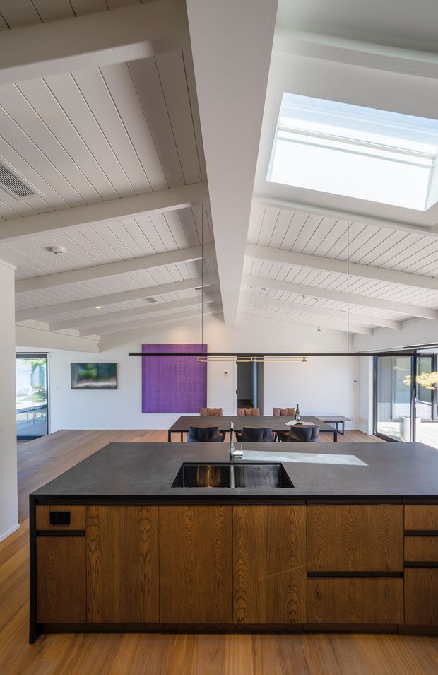 A spacious open-plan kitchen and dining area viewed from behind a large central island. The kitchen features dark charcoal cabinetry and benchtop, warm vertically grained timber joinery, an integrated black sink, and timber flooring. Beyond the island is a dining table with dark chairs, expansive glazing, and a purple feature artwork or wall panel. The room has a bright, airy character with exposed white-painted beams, a white-lined ceiling, and a large skylight.