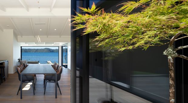A bright contemporary dining area opens directly toward expansive glazing and a blue waterfront view. The room features a long light-stone dining table with dark upholstered chairs, warm timber flooring, white painted walls and a coffered or battened ceiling. A dark-framed glazed opening and a sculptural potted tree create a strong transition between the interior and an adjacent terrace or courtyard.