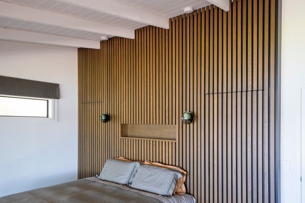 A contemporary bedroom featuring a full-height vertical timber-slat feature wall behind the bed, with an integrated recessed shelf and two dark spherical wall sconces. The room has a low-profile bed with neutral grey bedding, white walls, and a white panelled ceiling with exposed beam-like transitions. A narrow horizontal window with a dark roller blind provides natural light.