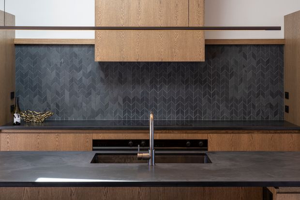 Contemporary kitchen interior featuring dark stone benchtops, warm timber cabinetry, a large central island with integrated sink and tap, and a dramatic dark geometric patterned tile splashback. Minimal black appliances and a slim horizontal timber detail reinforce the refined, modern composition.