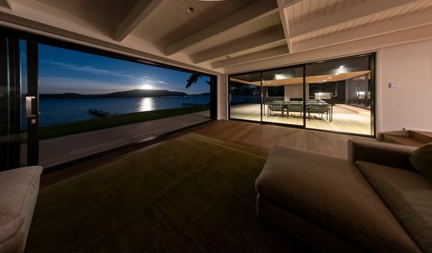 A dimly lit contemporary living room opening onto a waterfront deck, with expansive floor-to-ceiling glazing framing a moonlit bay and distant hills. The room features a large sectional sofa, dark timber flooring, a broad rug, and a distinctive white timber-lined ceiling with exposed beams. Through the glazed opening, a bright dining area with a table and chairs is visible.
