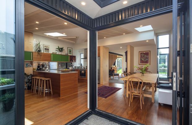 Interior view through dark-framed glazed doors into an open-plan kitchen and dining area. The kitchen features warm timber flooring, timber island cabinetry, green upper cabinet fronts, stainless-steel appliances and a skylit ceiling. The adjoining dining space has a timber table and bentwood chairs, with a dark timber-framed glazed opening and clerestory-style detailing creating a strong architectural feature.