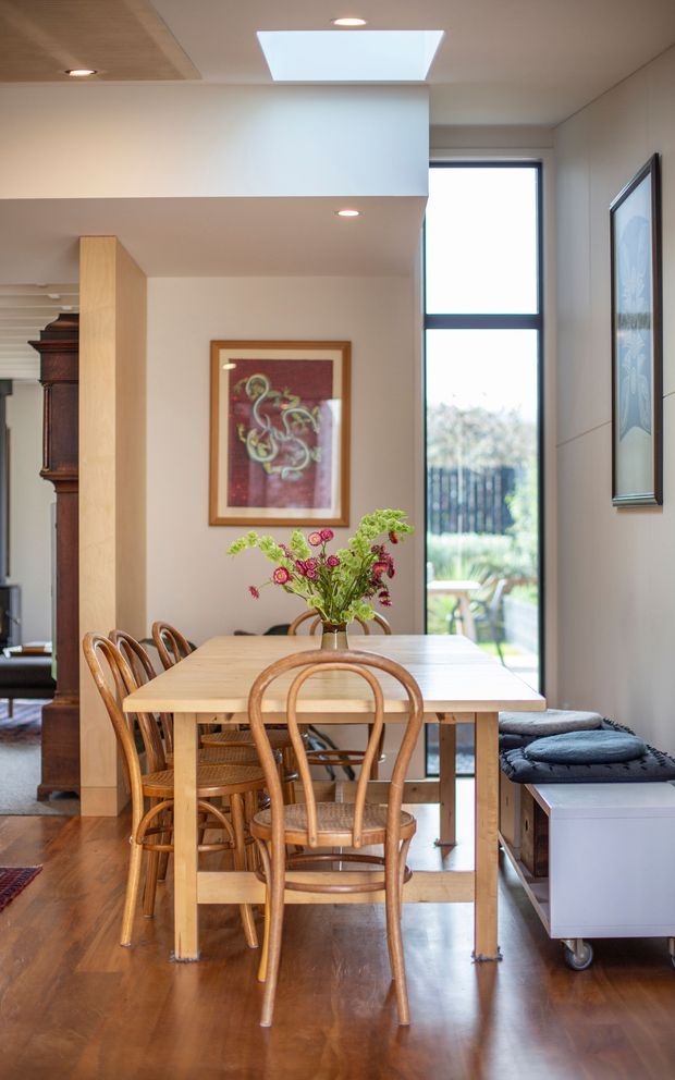 A bright, contemporary dining area with a long light-wood rectangular dining table surrounded by curved bentwood chairs. The room has warm timber flooring, white walls and ceiling, recessed downlights, a large skylight, and tall black-framed glazing overlooking greenery. A low upholstered bench or storage unit sits along the right wall, while artwork and a tall dark timber cabinet are visible in the adjoining space.
