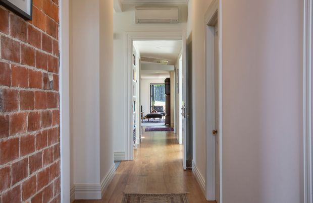 A long, bright residential hallway viewed down its length, with warm timber flooring, white walls and detailed white door and skirting trims. An exposed red brick feature wall runs along the left side, while multiple open doorways lead toward a furnished room and daylight at the far end. A wall-mounted split-system air conditioner is visible above the corridor.