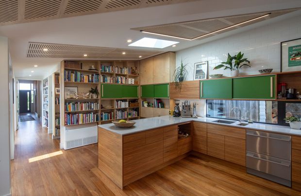 A warm contemporary kitchen with a timber island, extensive built-in shelving, green upper cabinetry, stainless-steel appliances, and a reflective tiled splashback. The open-plan space includes a visible hallway and integrated library joinery, with a slatted acoustic ceiling, skylight, and recessed lighting.