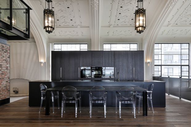 A dramatic open-plan kitchen and dining area in a heritage-style interior. A large dark kitchen island with integrated cabinetry and reflective splashback is paired with a long black dining table and transparent chairs. The room features an ornate white coffered ceiling, tall arched architectural openings, black-framed windows, pendant lanterns, exposed brickwork, and dark timber flooring.