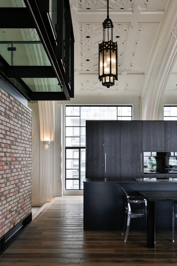 A sophisticated kitchen interior combining a dark, contemporary island and full-height black cabinetry with a historic decorative shell. The room features rich timber flooring, an exposed red-brick wall, large black-framed glazing, ornate white plaster ceiling panels and arches, and a prominent black metal pendant lantern. Clear acrylic dining or island stools add a modern contrast.