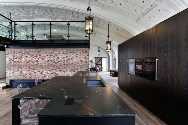 A spacious, sophisticated kitchen in a heritage-style interior, featuring a large dark stone island with an integrated sink and seating, dark timber cabinetry with built-in appliances, and a exposed brick feature wall. The room has an ornate white decorative ceiling, arched windows, black-framed glazing, and pendant lanterns, creating a refined industrial-contemporary atmosphere.