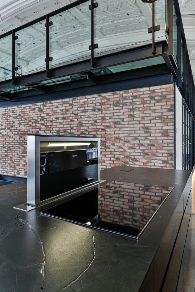 A dramatic commercial interior featuring a dark kitchen or service counter with a glossy black stone benchtop, integrated cooktop, and stainless-steel splashback or appliance surround. A large exposed red-and-grey brick wall forms the backdrop, while a black steel-framed glazed mezzanine with glass balustrades rises above. The ceiling has intricate exposed white structural detailing, creating an industrial warehouse-conversion atmosphere.