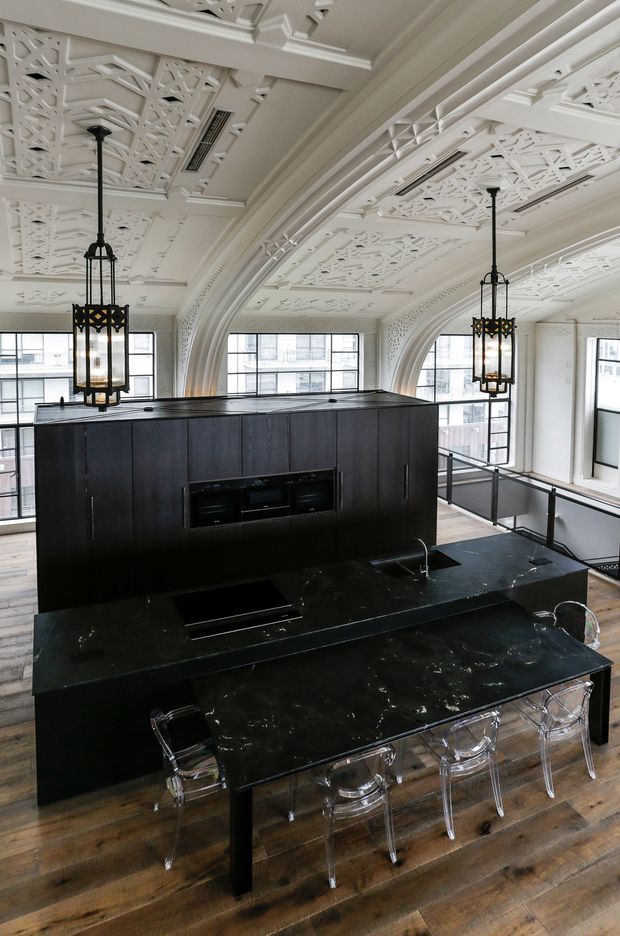 A dramatic contemporary kitchen in a grand heritage-style interior, featuring a large black stone island with integrated cooktop and sink, dark timber cabinetry, transparent bar stools, timber flooring, black-framed industrial-style windows, and ornate white decorative ceiling panels with curved architectural arches. Oversized black lantern pendants add contrast.