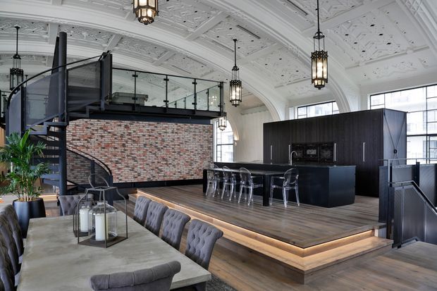A spacious, high-ceilinged loft-style interior combining a dining area, raised kitchen platform, and mezzanine. The room features ornate white decorative ceiling panels, exposed brickwork, dark cabinetry, black metal stair and balustrade detailing, timber flooring, and industrial pendant lights. A large dining table with upholstered seating occupies the foreground, while a compact black kitchen and transparent bar stools sit on the raised timber platform.