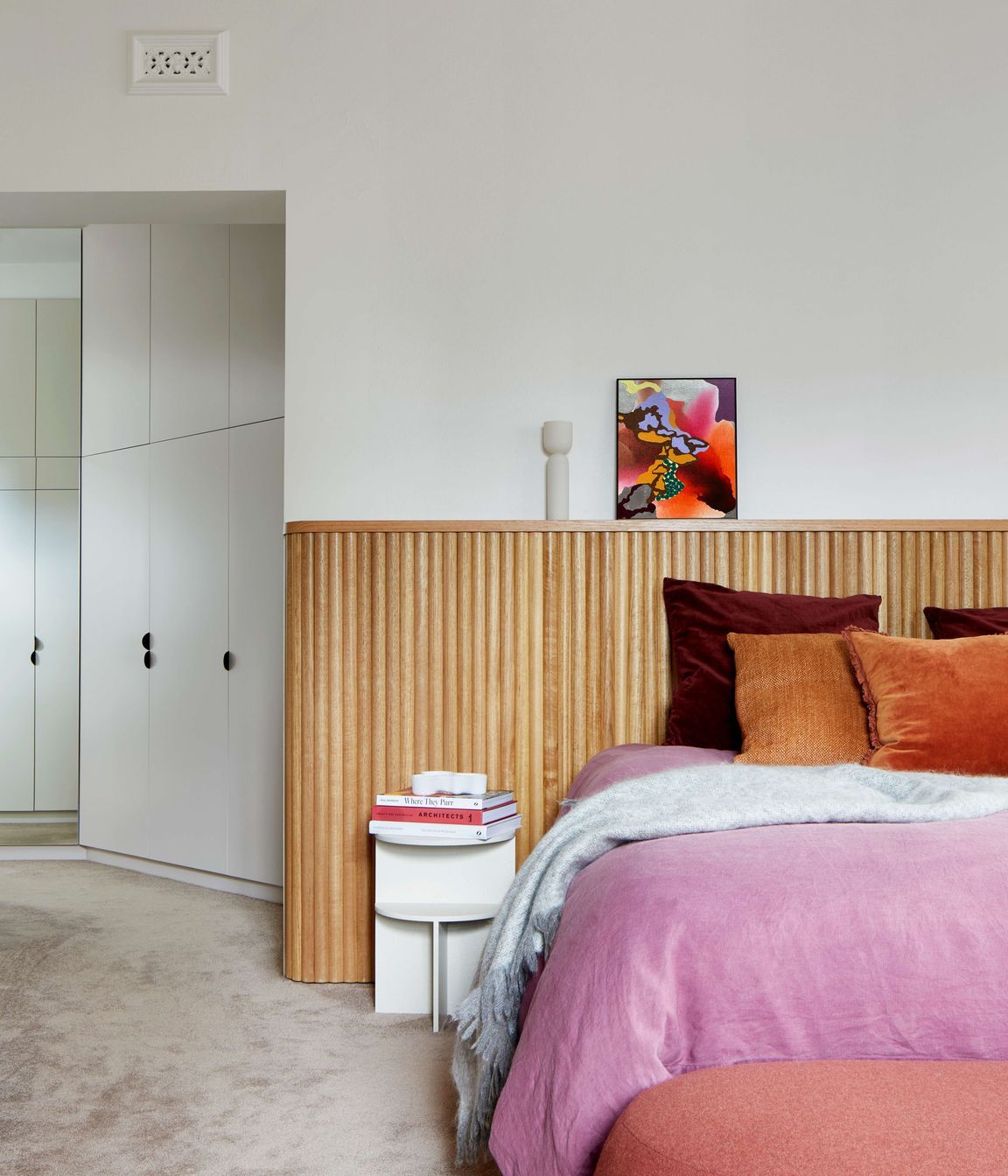 Master bedroom with headboard created with tasmanian oak wood dowels for a highly finished art deco minimalist feel. Cushions and bedding provide pops of colour in warm pink and jeweled colour oranges and burgundies. Pale pink  coloured carpet from Edel Carpets. Home located in Melbourne. See more from our Arch Deco Project.